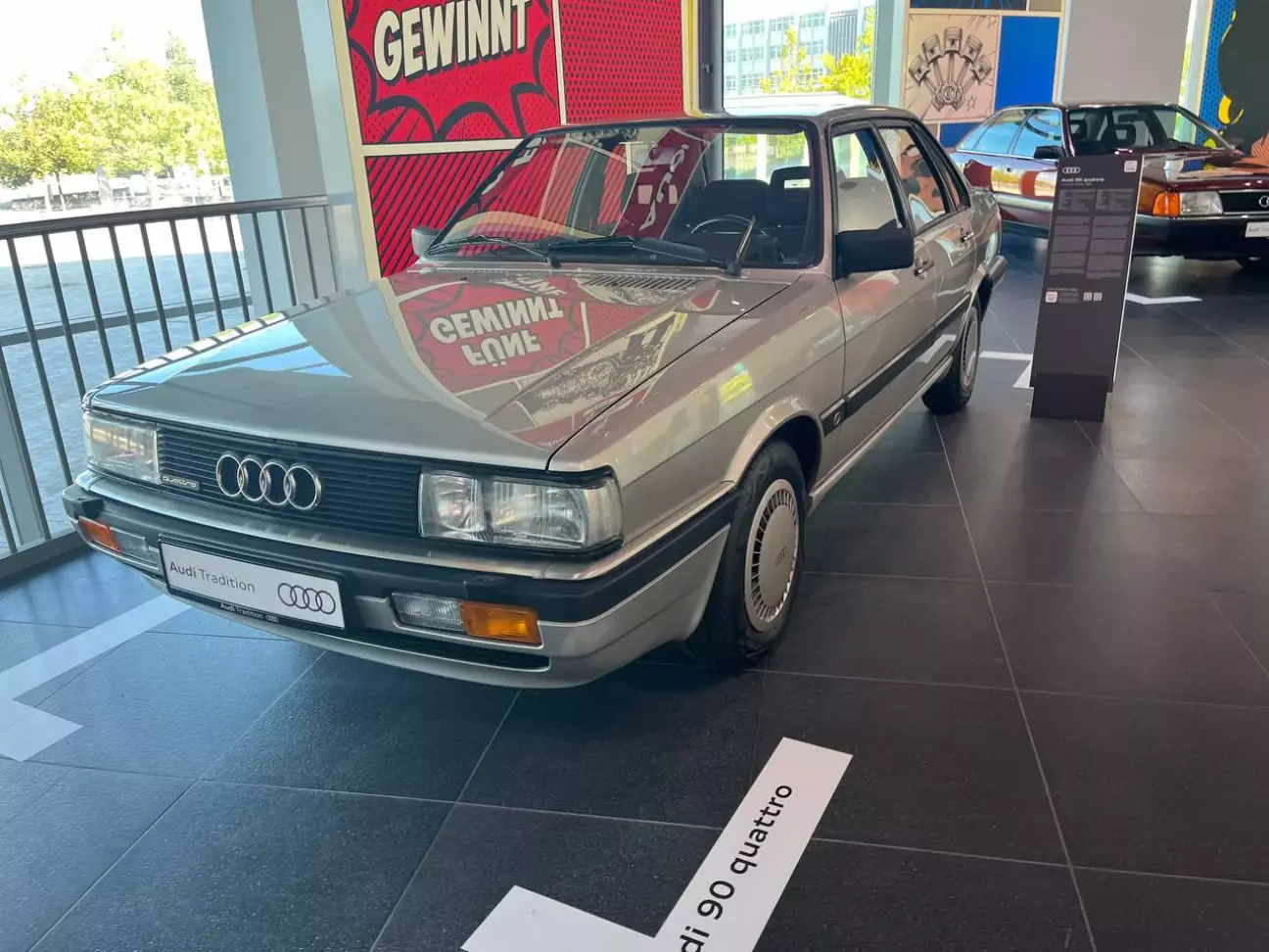Покоління B2, Audi 90 — з 1984 року Двигуни (версії моделі) рядні 5-циліндрові 2,0 л і 2,2 л Потужність (версії моделі) 115–136 к.с. (85–100 кВт) 2,2 л з каталізатором (quattro) 120 к.с. (88 кВт) Привід постійний повний quattro Кінець виробництва B2 серпень 1986 року Audi 90 з'явився в 1984 році разом з оновленням Audi 80 другого покоління (B2). Це була більш розкішна «сестра» вісімдесятки, і, як зазначає німецька Wikipedia, відомі п'ятициліндрові мотори відтепер залишили саме для цих краще оснащених версій. Від Audi 80 модель відрізнялася широкими фарами, якіснішою оббивкою салону, вставками з нержавної сталі в захисних молдингах, а також бамперами й накладками порогів від Audi Coupé GT. Експонат — сріблястий седан із шильдиком quattro на решітці радіатора, ковпаками на колесах і оранжевими покажчиками повороту в бампері. На підлозі перед ним — напис «Audi 90 quattro». Під капотом Audi 90 стояли рядні п'ятициліндрові мотори. За даними англійської Wikipedia, гама включала 2,0-літровий агрегат на 115 к.с. і 2,2-літровий на 136 к.с.; версії з каталізатором 2,2 л розвивали 115 к.с. з переднім приводом і 120 к.с. — у модифікації quattro. Якою саме версією є експонат, табличка з відстані не дозволяє прочитати, тому ці цифри варто сприймати як характеристики моделі загалом. Важливіше інше: Audi 90 закріпив за п'ятьма циліндрами статус ознаки старших версій компактної моделі, а не лише великих седанів Audi 100 і 200. Повний привід quattro до того часу вже встиг стати легендою. У березні 1980 року Audi показала в Женеві однойменне купе з турбованим п'ятициліндровим мотором і постійним повним приводом, а невдовзі quattro почав вигравати чемпіонати світу з ралі. Наступним логічним кроком було перенести цю технологію у звичайні седани. Ще до появи Audi 90 повний привід отримав Audi 80: за даними Wikipedia, у 1982–1984 роках пропонувався Audi 80 quattro з п'ятициліндровим мотором 2144 см³ і 136 к.с. Audi 90 був так само доступний у версії quattro, і саме такий автомобіль стоїть на виставці. Виробництво B2 завершилося в серпні 1986 року, а вже наступна генерація B3 отримала власний Audi 90 з новим дизайном. Однак лінія, яку започаткував цей сріблястий седан, тривала довго. На платформі середнього класу пізніше з'явилися Audi Coupé S2 та легендарний Avant RS2 — обидва з п'ятициліндровими турбомоторами, і обидва стоять на цій самій виставці. За даними Audi, п'ятициліндрові мотори залишили сегмент B лише в 1994 році, коли на зміну Audi 80 прийшов A4 (B5), а V6 поступово витіснили їх з гами. Тому Audi 90 quattro — важливий проміжний етап: від великих седанів до компактних спортивних машин.