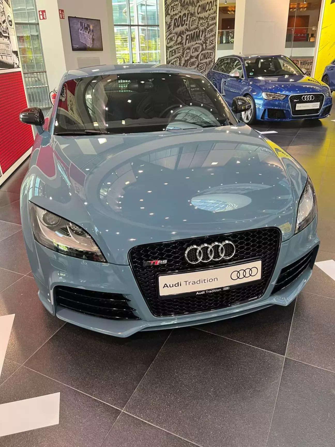 Двигун 2,5 TFSI, рядний 5-циліндровий, турбонаддув, безпосереднє впорскування, 2480 см³ Потужність (TT RS 2009) 250 кВт (340 к.с.) при 5400–6500 об/хв Крутний момент (TT RS 2009) 450 Нм при 1600–5300 об/хв Трансмісія (на старті продажів) 6-ступінчаста механічна, постійний повний привід quattro 0–100 км/год (купе) 4,6 с Максимальна швидкість 250 км/год (280 км/год опційно) TT RS plus (2012) 265 кВт (360 к.с.), 465 Нм Назва виставки «1-2-4-5-3» — це порядок роботи циліндрів п'ятициліндрового мотора Audi, і світлий сіро-блакитний TT RS стоїть тут не випадково. У 1997 році з виробництва пішли останні «п'ятірки» першої епохи — 2,5-літровий TDI в A6 і 2,2-літровий 20V turbo в S6, — і на дванадцять років цей формат зник із гами. Повернення відбулося 2009-го: на Женевському автосалоні дебютував TT RS із поперечно встановленим 2,5-літровим TFSI. Модель підготувала quattro GmbH — той самий підрозділ, що створював RS 4 і RS 6, — і саме з неї почалася нова біографія п'ятициліндрового двигуна, яка триває й досі. Двигун 2,5 TFSI створили на основі концернового агрегата EA 855, але з посиленим блоком циліндрів. Робочий об'єм — 2480 см³ (діаметр циліндра 82,5 мм, хід поршня 92,8 мм), ступінь стиснення 10,0:1, надлишковий тиск наддуву — до 1,2 бара. Безпосереднє впорскування й інтеркулер дозволили отримати 250 кВт (340 к.с.) у діапазоні 5400–6500 об/хв, а пікові 450 Нм доступні від 1600 до 5300 об/хв. Сам мотор важить 183 кг. Порядок роботи 1-2-4-5-3 чергує запалювання в сусідніх і віддалених циліндрах — недарма на одному з панно виставки написано: «It's not just power. It's rhythm». Купе отримало шестиступінчасту механічну коробку передач і постійний повний привід quattro з електронно керованою багатодисковою муфтою з гідравлічним приводом. Розгін 0–100 км/год займав 4,6 с, максимальну швидкість обмежували на 250 км/год, а за бажанням клієнта межу піднімали до 280 км/год. Кнопка Sport на центральному тунелі відкривала заслінку у вихлопній системі й робила реакцію на педаль газу гострішою. 2012 року з'явилася версія TT RS plus: 265 кВт (360 к.с.) і 465 Нм. А сам двигун у 2010–2018 роках дев'ять разів поспіль перемагав у класі 2–2,5 л на конкурсі International Engine of the Year. Експонат — купе світлого сіро-блакитного кольору; на стенді поруч зазначено просто «Audi TT RS» і рік 2012, тож про конкретну версію чи комплектацію саме цього автомобіля ми судити не беремося. Чорна стільникова решітка радіатора з шильдиком TT RS, табличка Audi Tradition замість номерного знака і поп-арт-оформлення зали з вигуками «Fünf!» кількома мовами створюють правильний настрій. Це місток між культовими п'ятициліндровими Audi 1980-х і сучасними RS 3, які стоять буквально за кілька кроків. Якщо шукати в експозиції точку, де стара легенда знайшла друге дихання, — вона саме тут, біля цього купе.